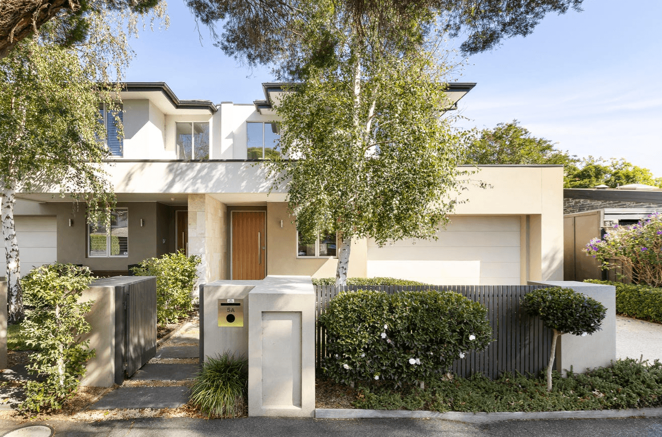 Modern two-story beige house with a wooden front door, gated entry, and landscaped front yard with trees and shrubs.