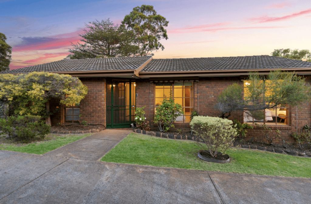 Brick ranch-style house with a tiled gray roof at sunset, front yard shrubs, and a paved driveway.