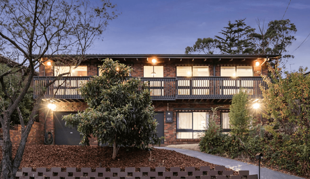 Two-story brick townhouse with a wooden second-floor balcony, front yard lighting, and trees at dusk.