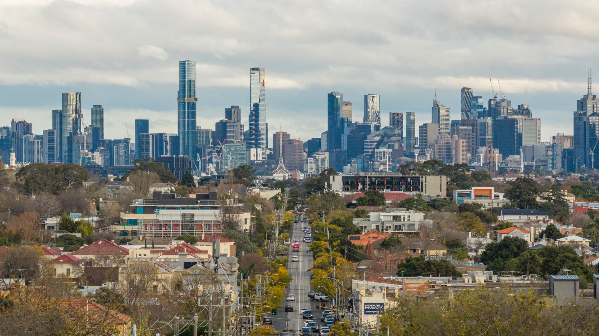 Wide view of a modern city skyline with tall glass skyscrapers rising behind a tree-lined residential street and a central road in the foreground.