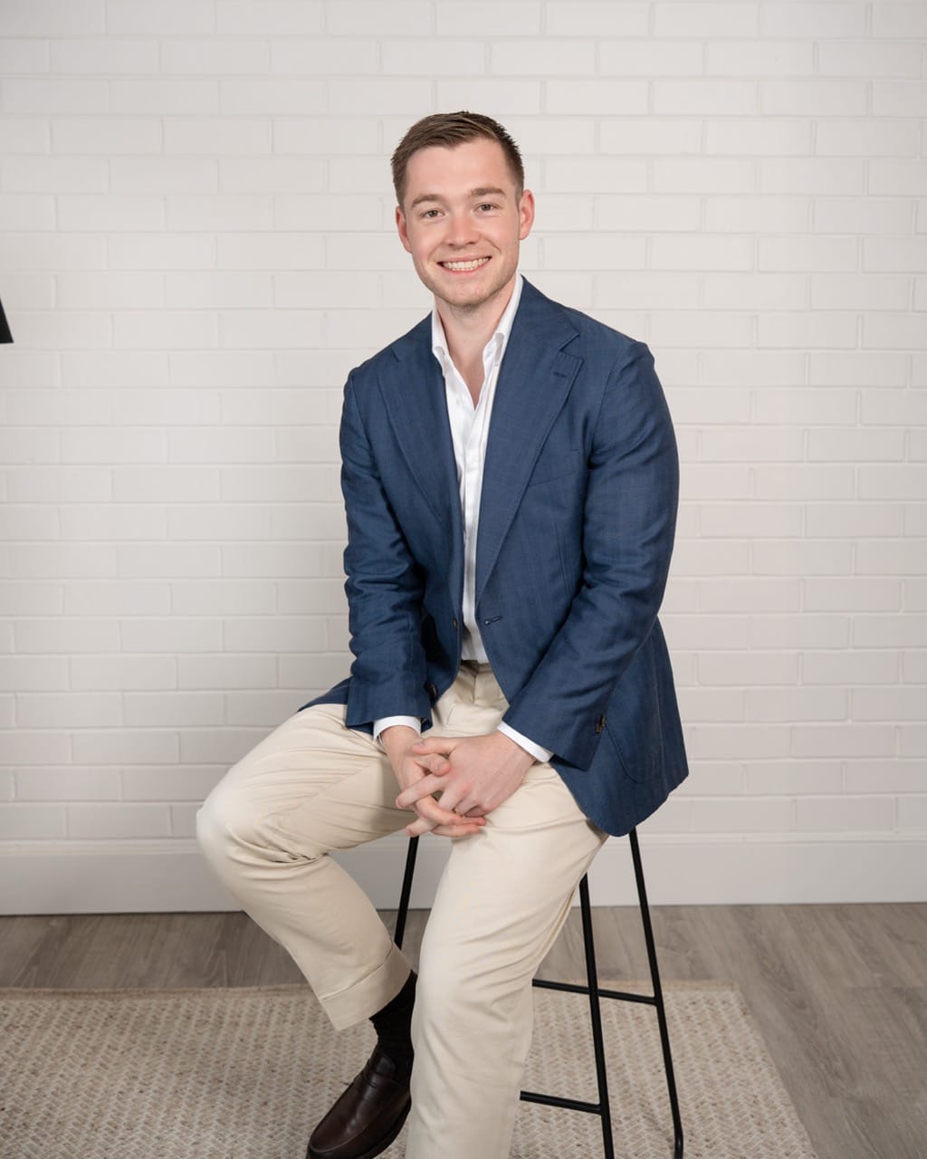 Smiling man in a blue blazer and white shirt sitting on a black stool against a white brick wall.