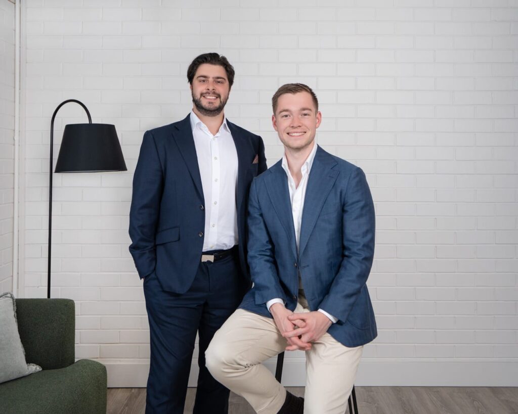 Two men in business attire posing together in a modern room with a white brick wall behind them.