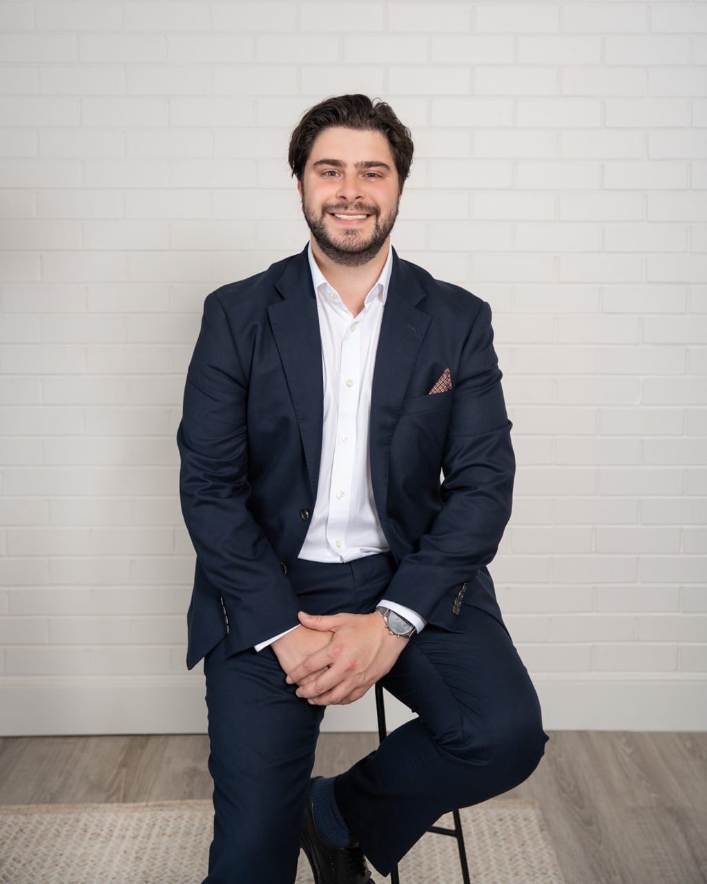 Smiling man in a navy suit and white shirt sits on a stool against a white brick wall, hands folded in his lap.