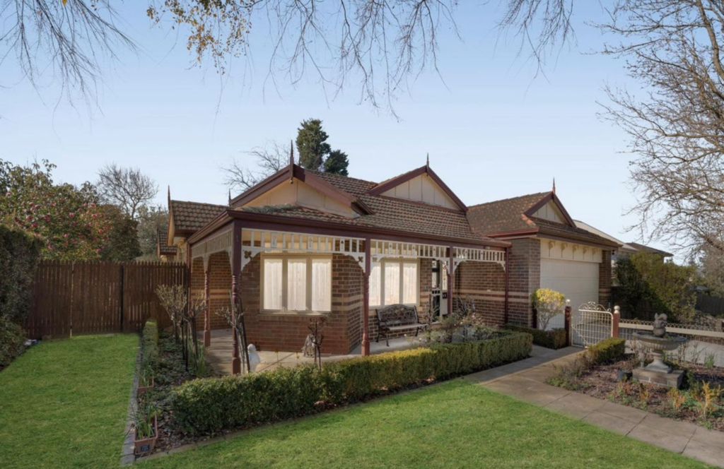 Brick bungalow with a red-tiled roof, white trim and a covered porch, set in a manicured front yard under a clear blue sky.